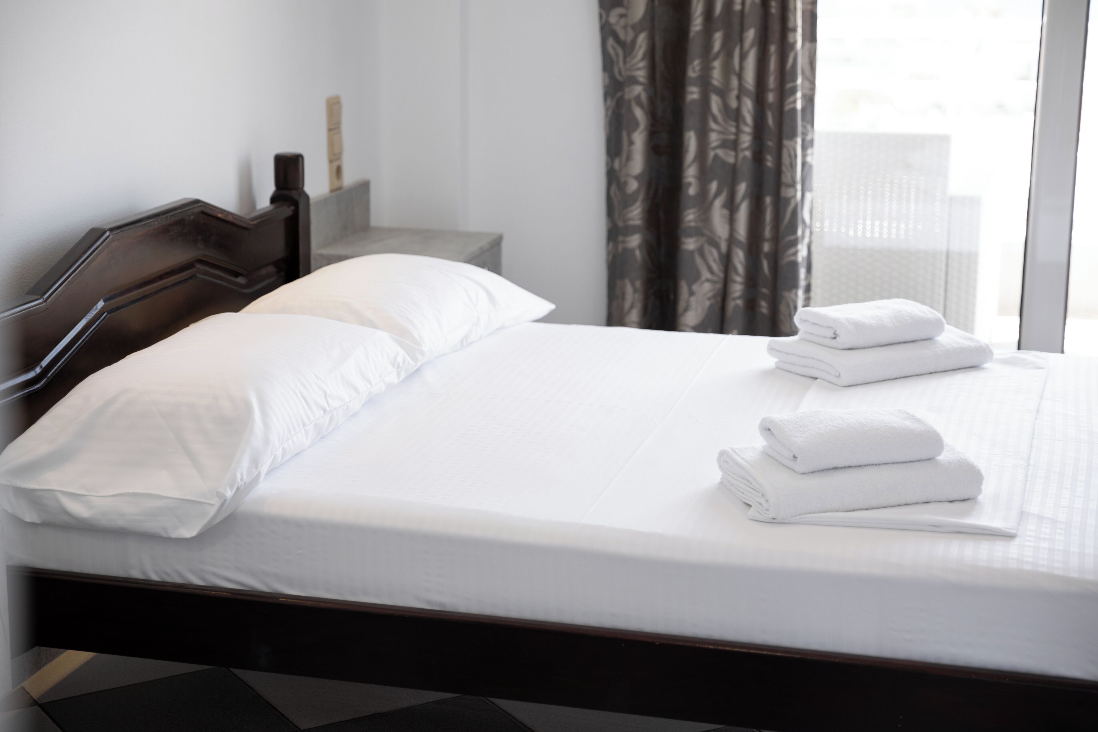Un lit bien fait, avec du linge de lit blanc et des serviettes pliées, une tête de lit en bois, une table de chevet avec une lampe, et une vue sur un bâtiment à travers une fenêtre avec des rideaux.