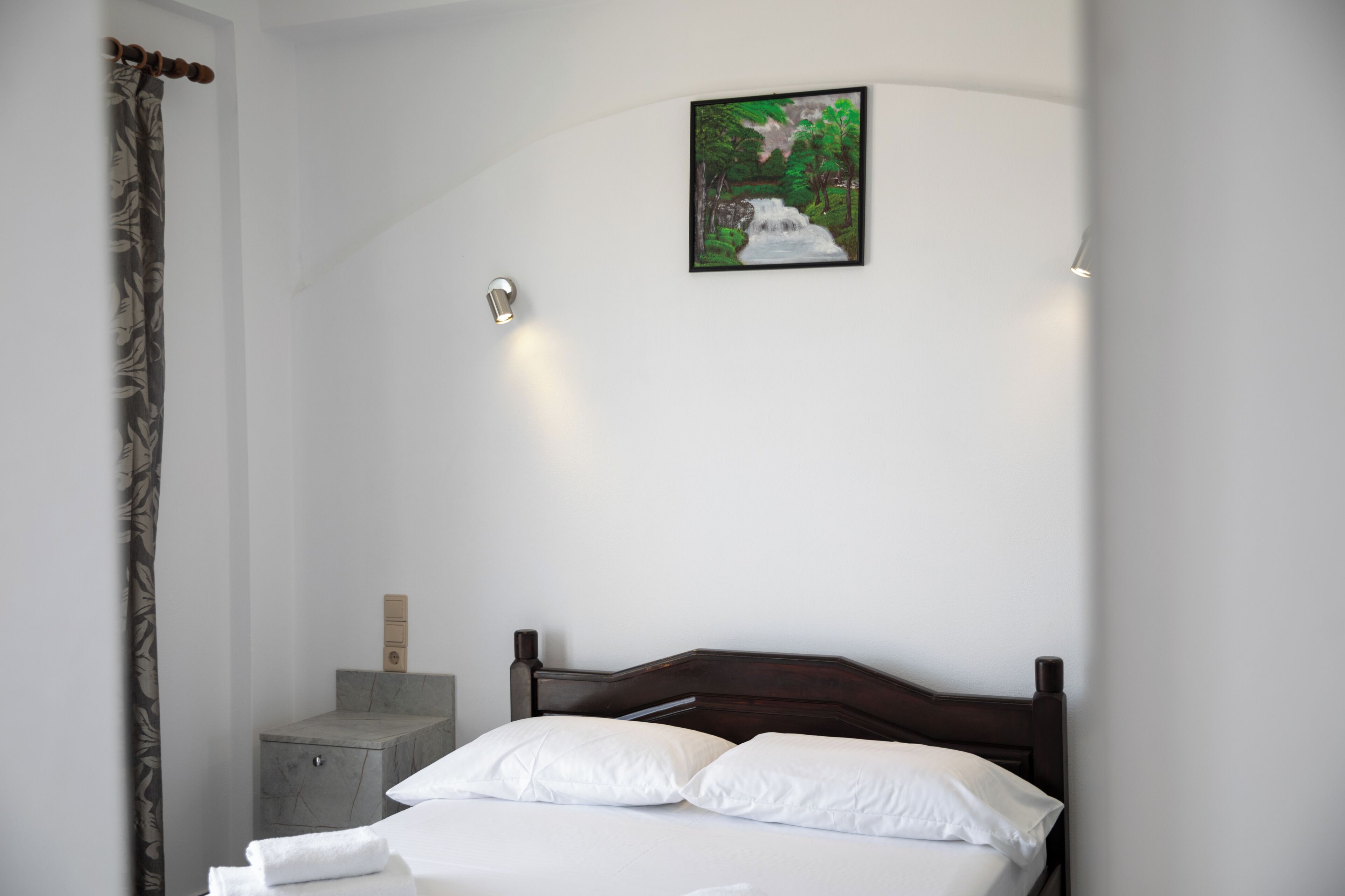 Een net opgemaakt bed met witte lakens, een houten hoofdbord en een lijst met een schilderij van een waterval aan de muur.