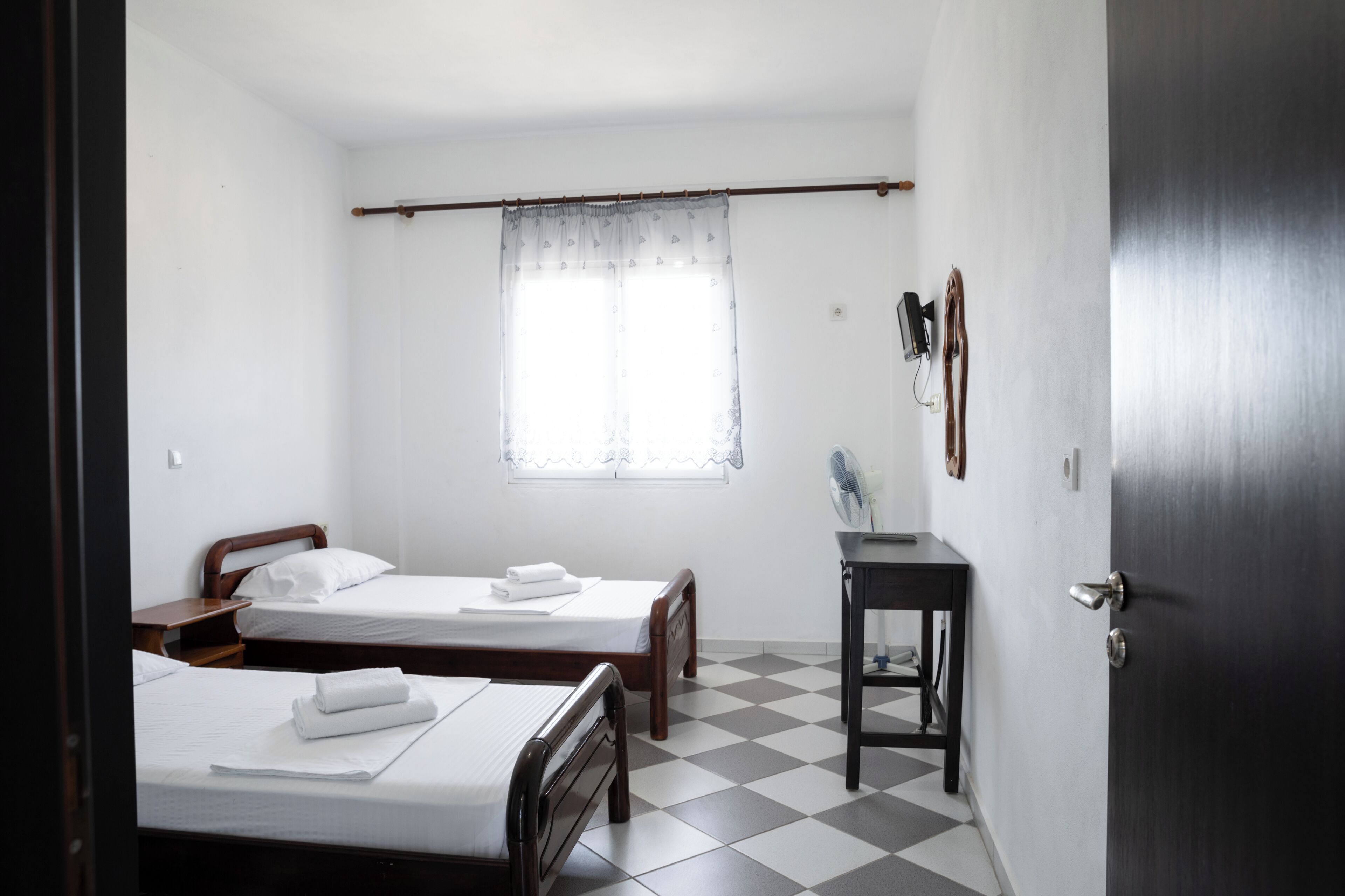 Una habitación con dos camas individuales, una mesita pequeña, una ventana con cortinas translúcidas y una puerta.