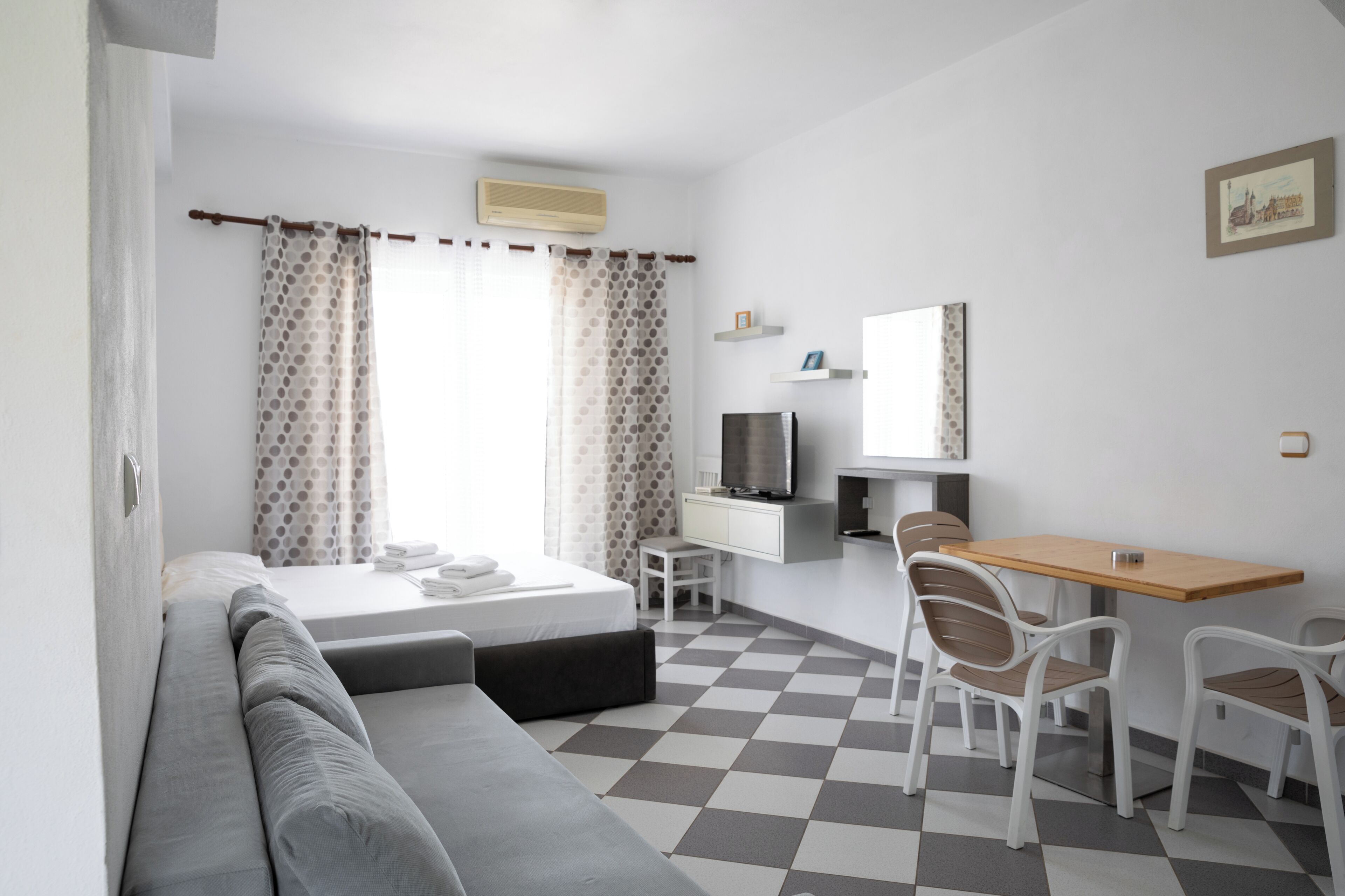 A hotel room with a bed, a sofa, a desk with chairs, a television, and a checkered floor.