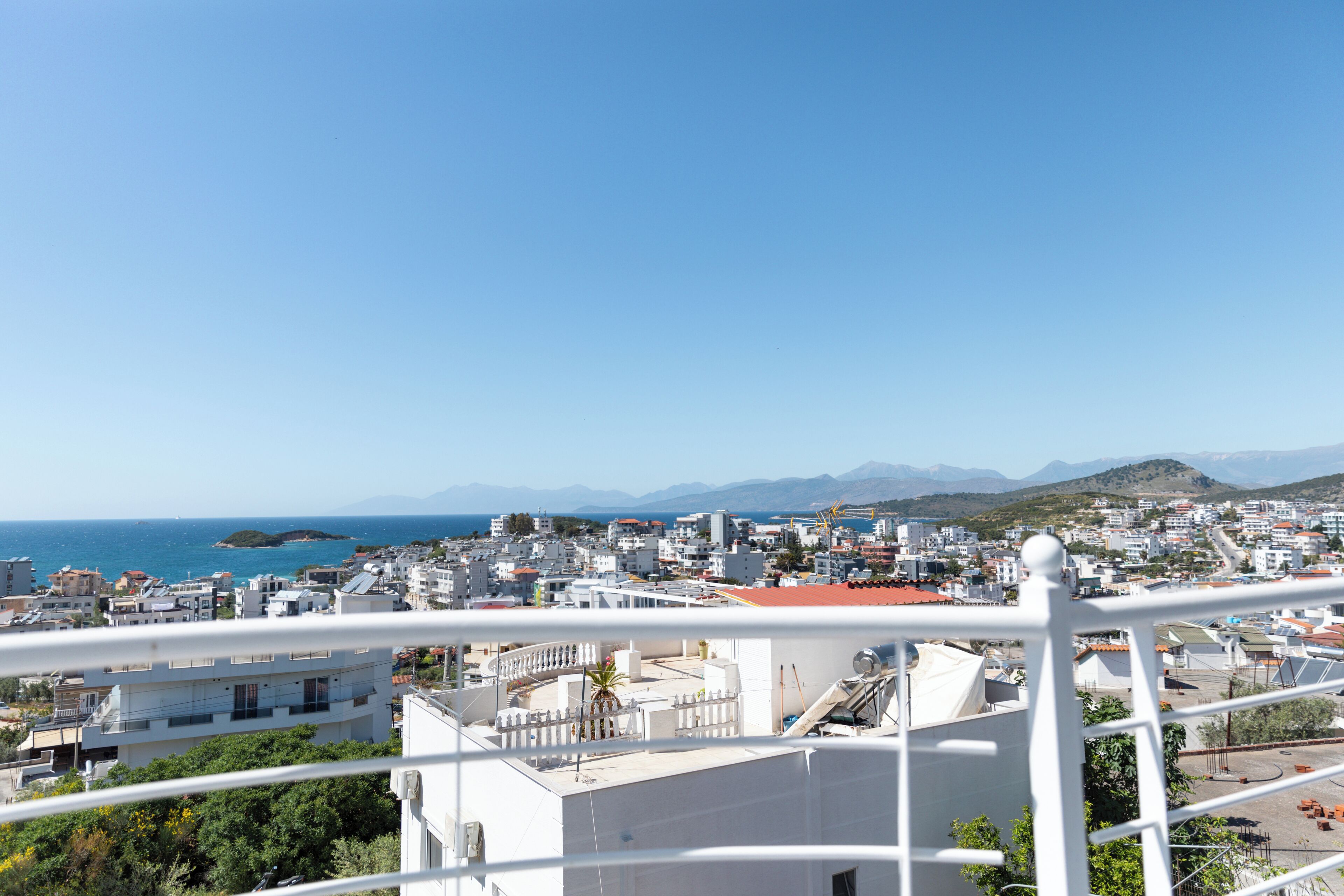 A coastal cityscape with white buildings, a clear blue sky, and a view of the sea and mountains in the distance.