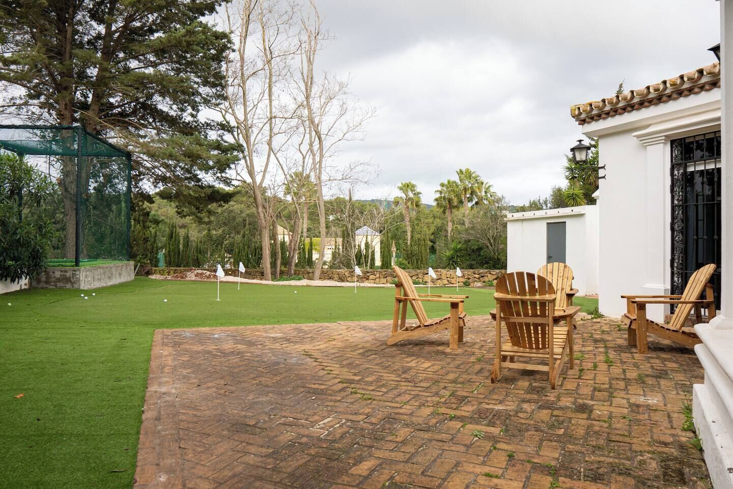 A patio with brick flooring, wooden chairs, and a lawn with a golf hole.