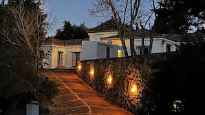 Uma trilha conduz a um edifício com parede de pedra e arandelas iluminadas.