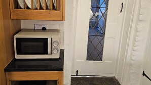 A compact kitchen with a microwave, wooden shelves with dishes, wicker baskets, and a view through a door with a diamond-patterned window. - Beautiful historic cottage pets welcome (Birchington-on-Sea)
