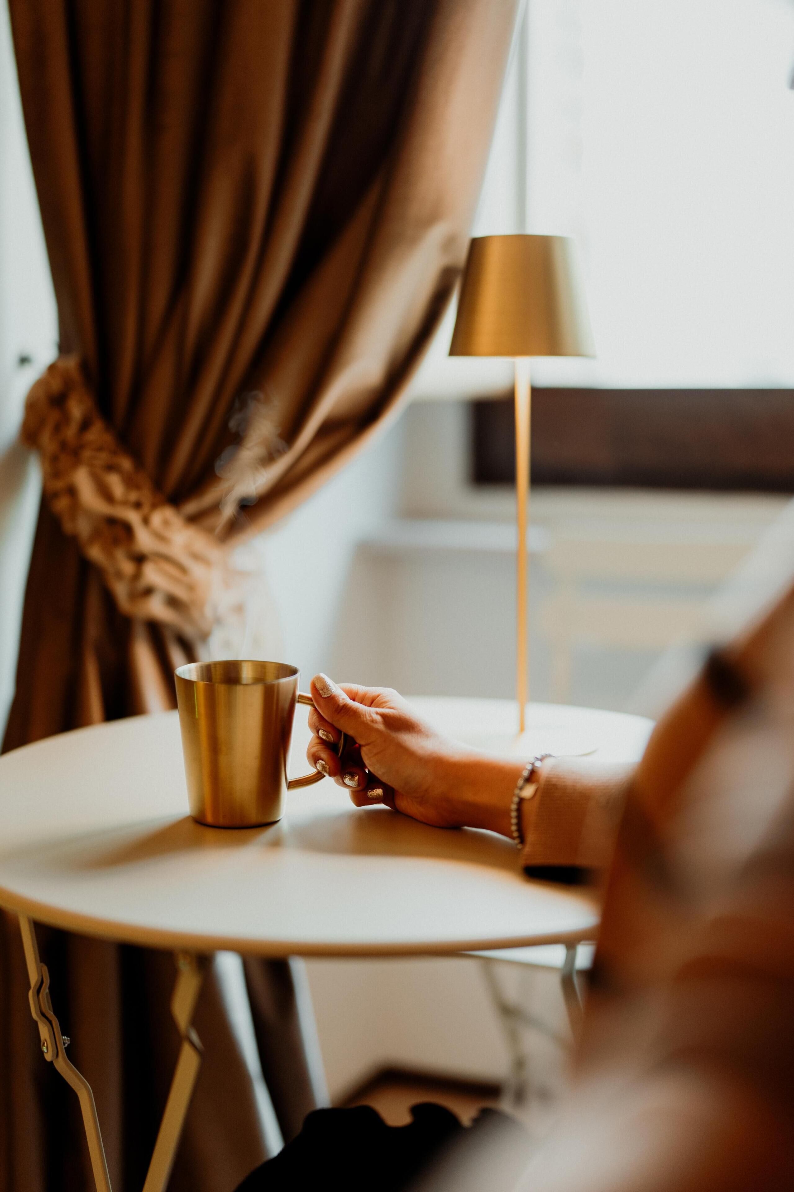 Iemand zit aan een ronde tafel met een goudkleurig kopje, een lamp en gordijnen op de achtergrond.