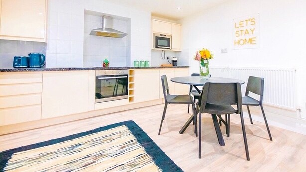 Une cuisine moderne avec des meubles blancs, un four à micro-ondes et une cuisinière. On y trouve une table à manger avec quatre chaises et un vase rempli de fleurs. Une pancarte sur le mur porte l'inscription : « RESTONS À LA MAISON ».