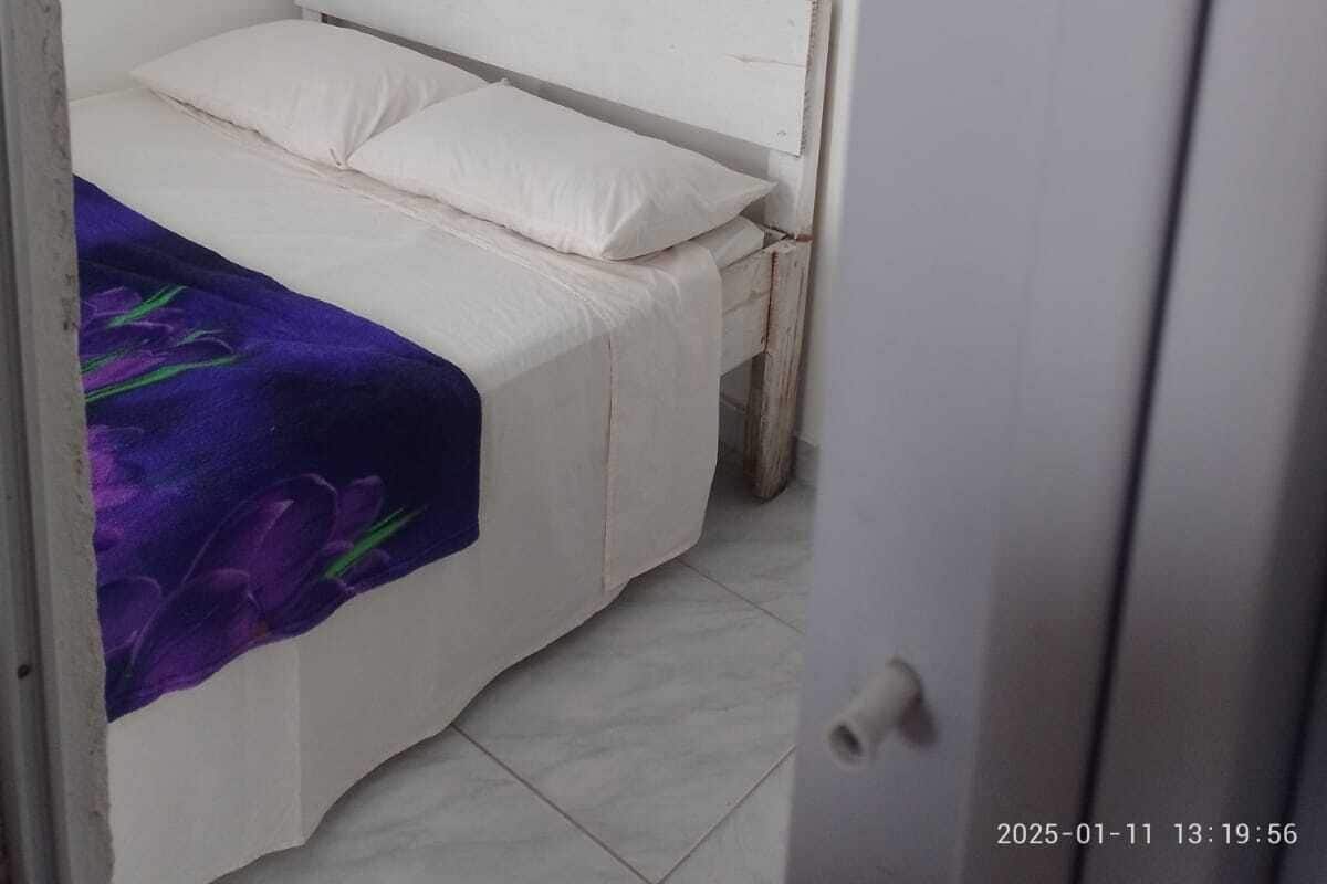 A small, single-bed room with a white bed and a purple blanket. There is a white air conditioning unit mounted on the wall above the bed.