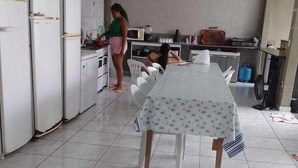 Une cuisine avec des appareils électroménagers blancs, une table à manger recouverte d’une nappe et un sol carrelé.