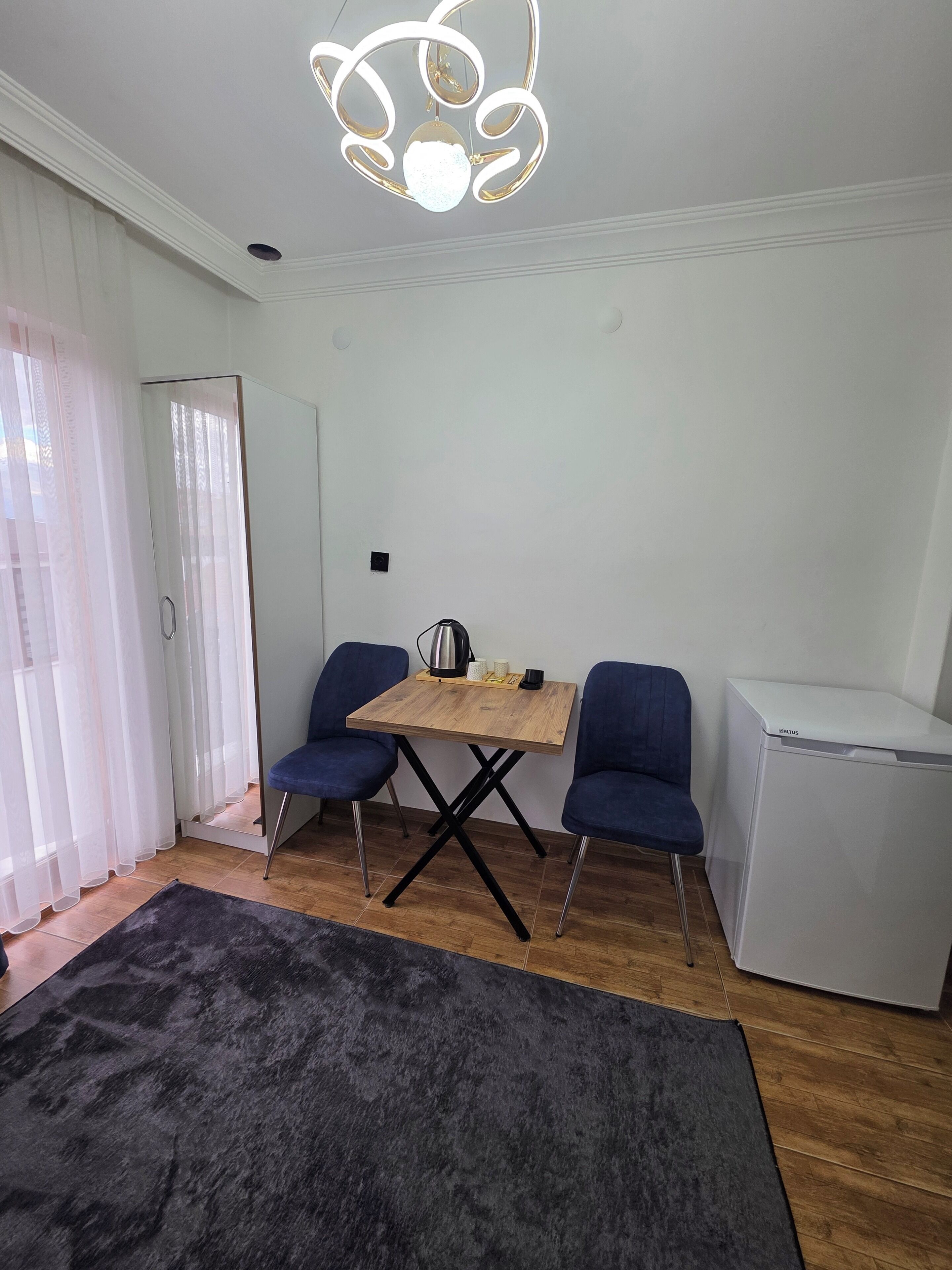 Una habitación moderna con una mesa de madera, dos sillas azules, un tapete negro, un refrigerador blanco y una lámpara de techo original.