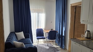 Una sala de estar moderna con un sofá azul oscuro, una pequeña mesa de comedor con dos sillas y una cocina con armarios y una tetera.