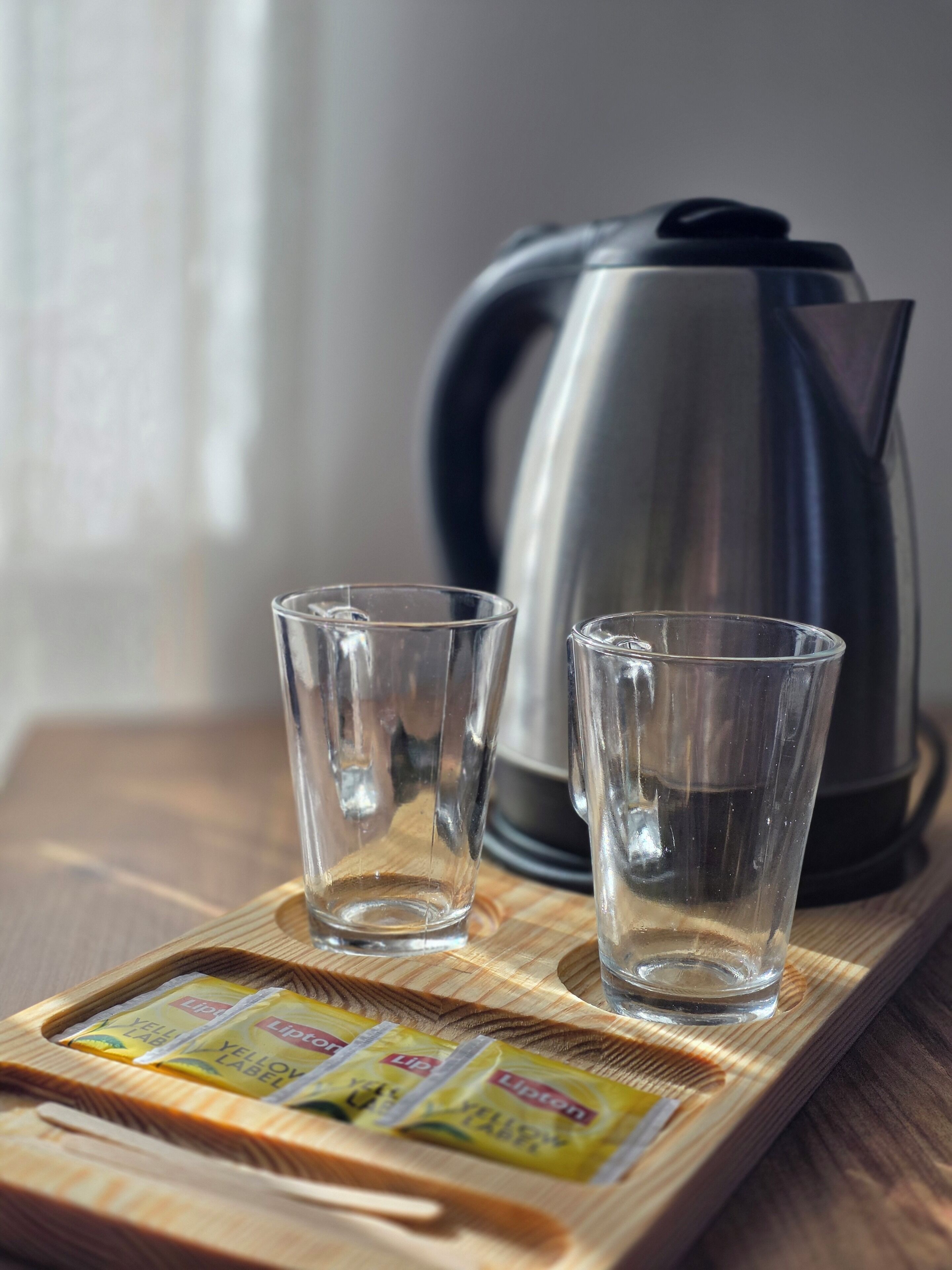 Two clear glasses on a wooden tray with Lipton tea packets, a kettle, and a window in the background.