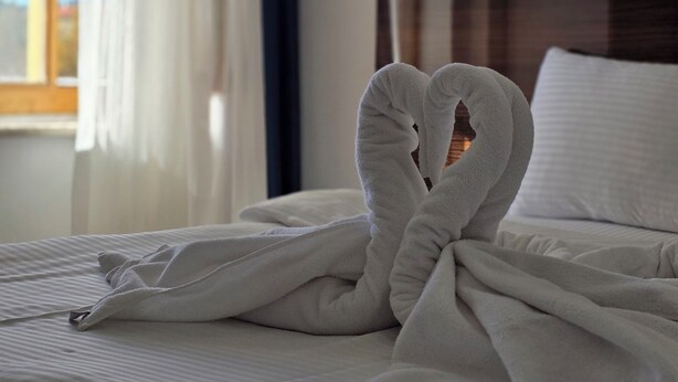 Una cama bien hecha con una toalla doblada en forma de corazón en el centro, un cabecero de madera y una ventana con cortinas.