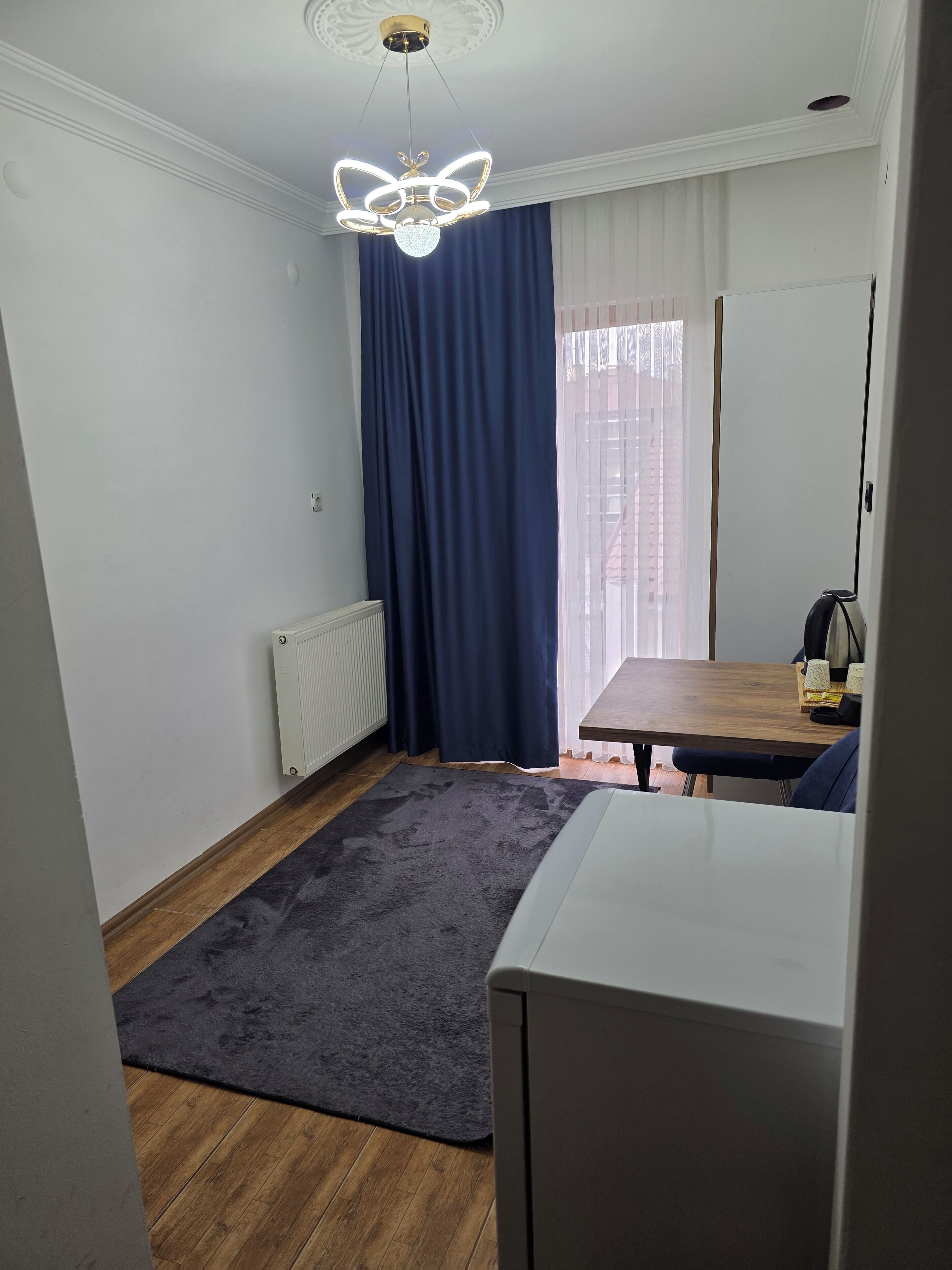 A compact room with a wooden floor, a dark blue curtain, a white radiator, a small table with a chair, and a modern light fixture.
