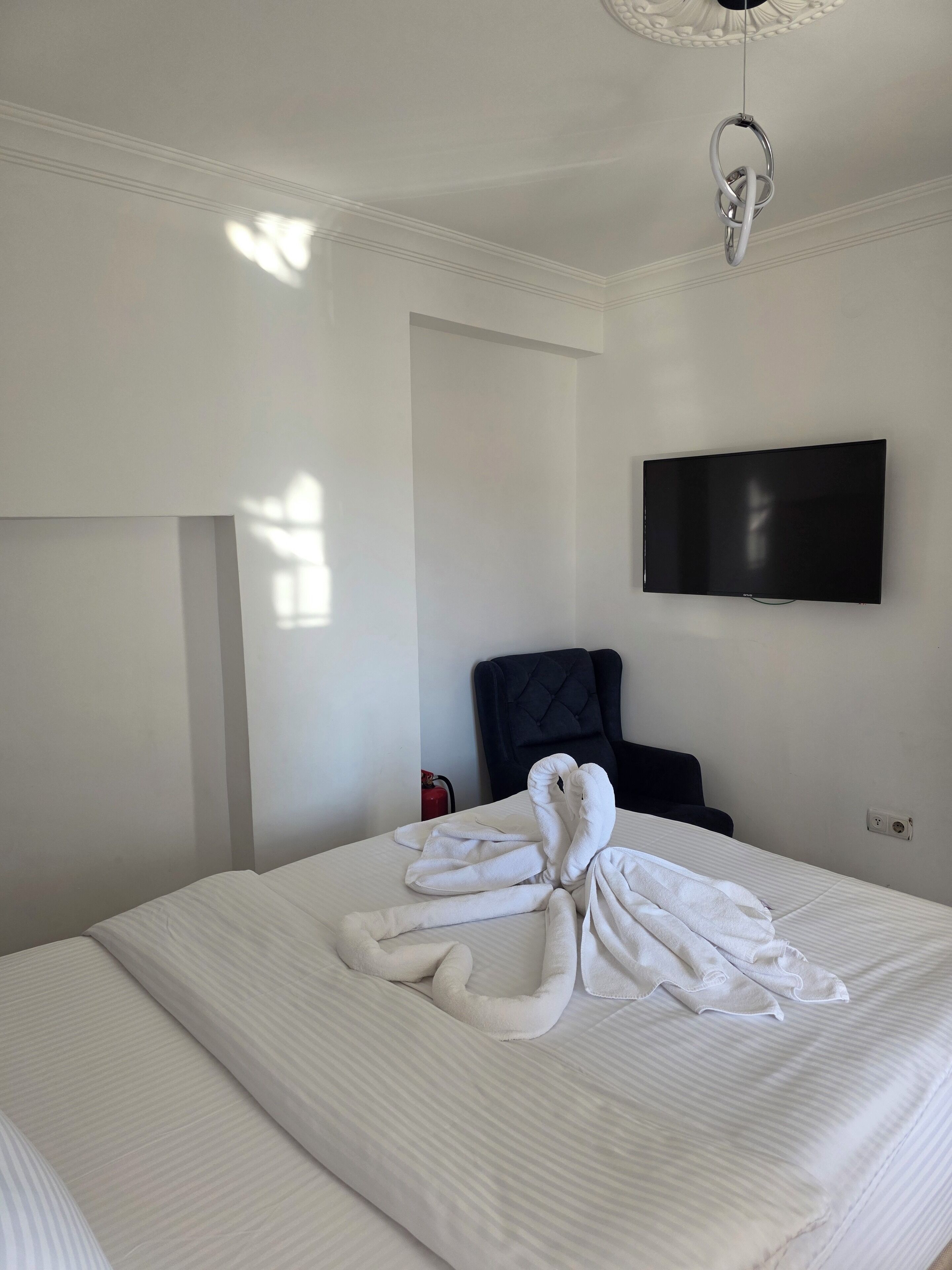 A hotel room with a neatly made bed, a flat-screen TV mounted on the wall, a black upholstered chair, and a ceiling light fixture.