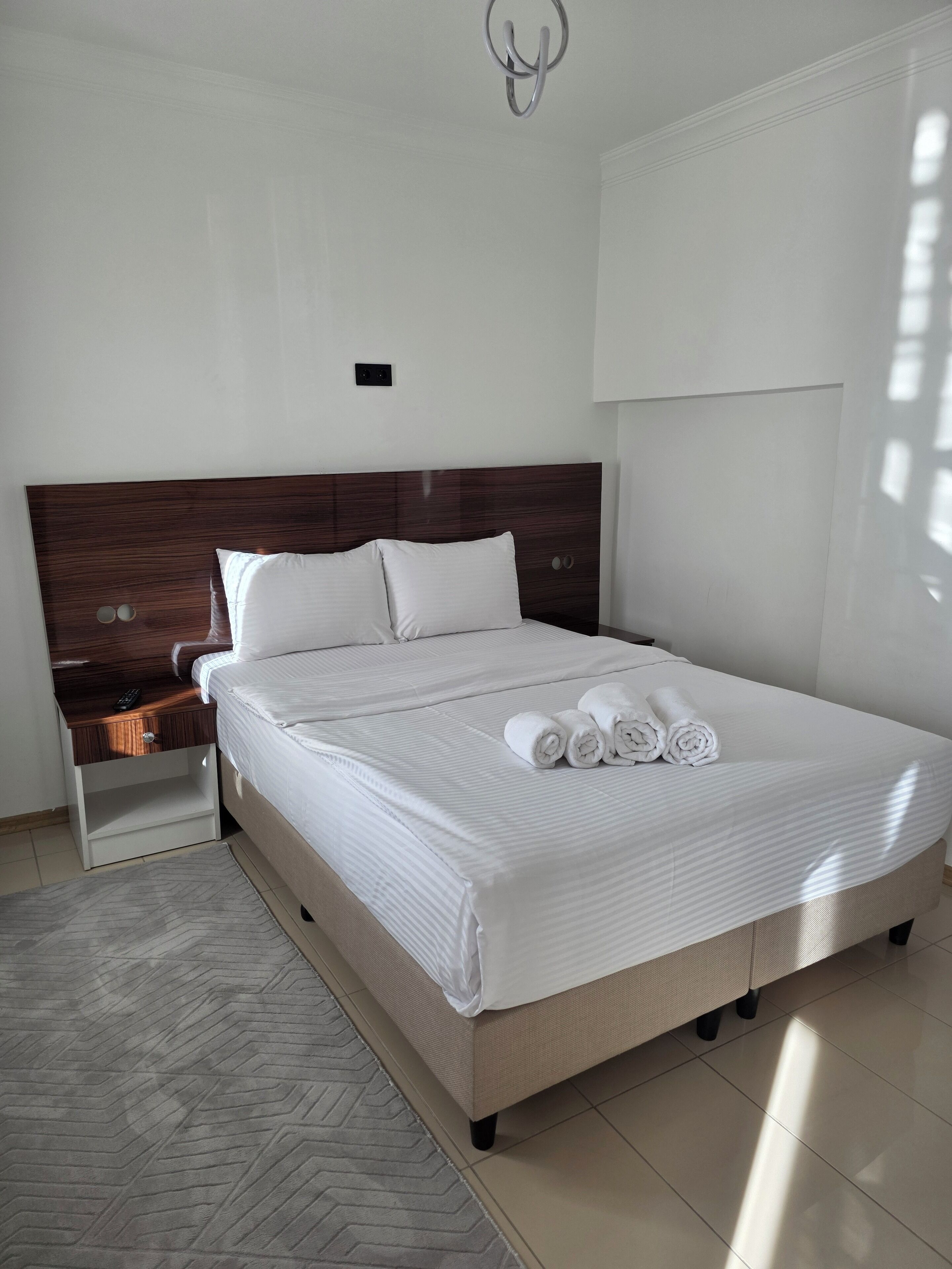 A neatly made bed with white linens and rolled towels, a wooden headboard, a bedside table with a remote control, and a patterned rug.