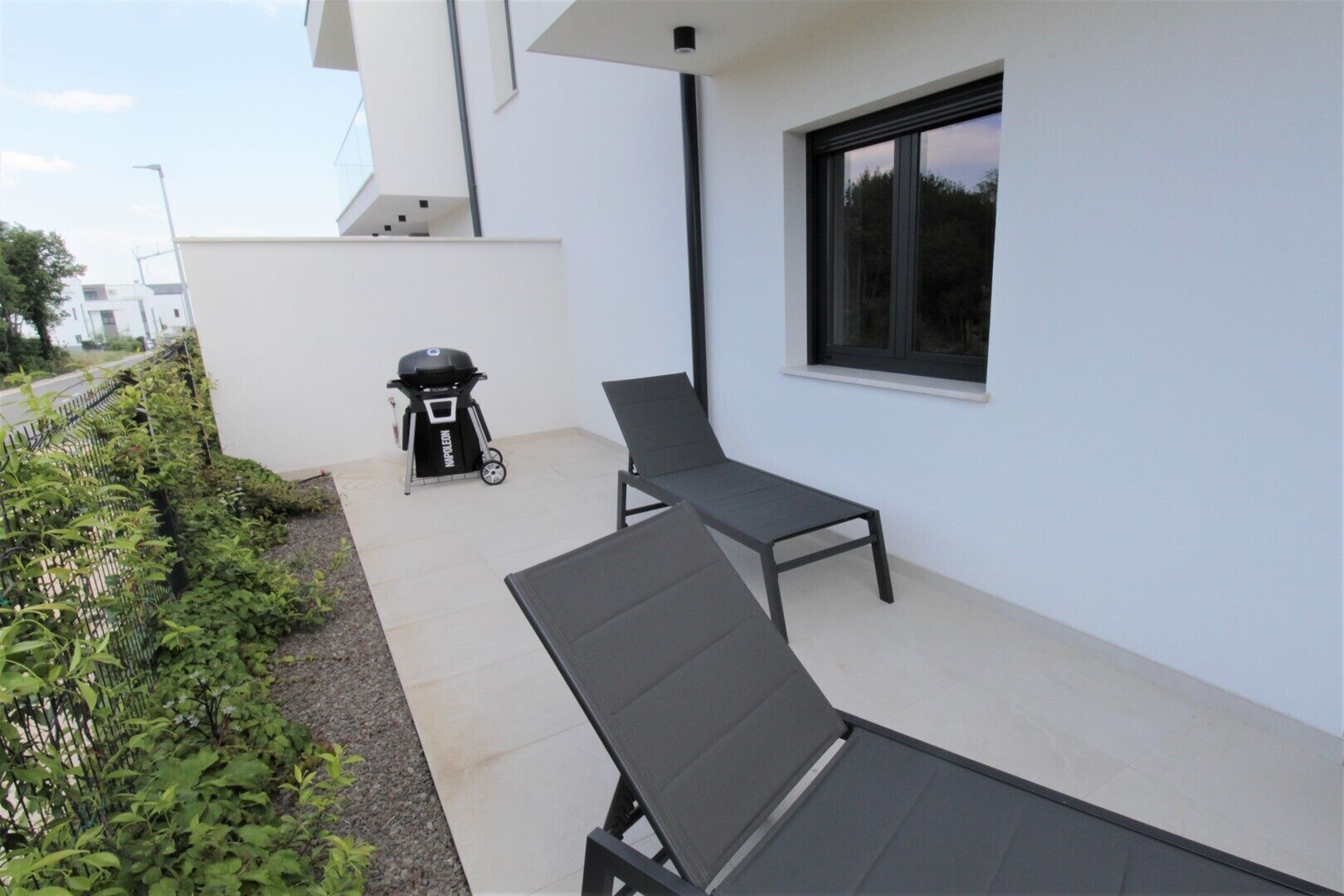 A patio with two lounge chairs, a barbecue grill, and a window overlooking greenery.
