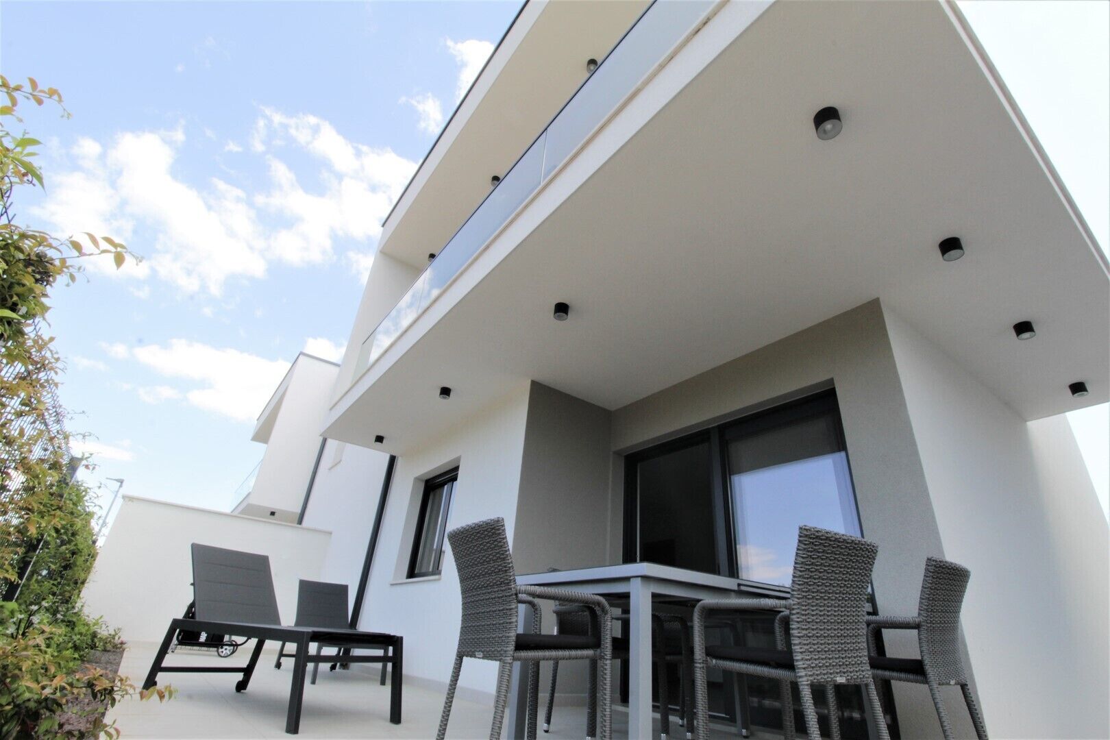 Une terrasse extérieure moderne, avec une table et des chaises, une grande fenêtre et une façade blanche.