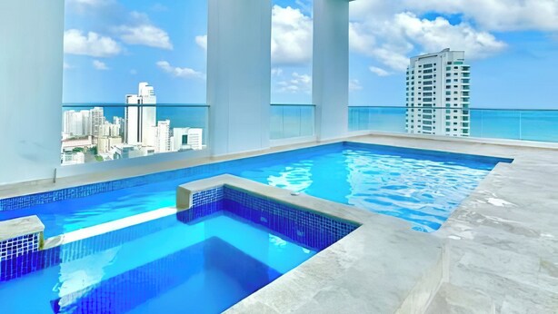 Une piscine intérieure moderne, avec une eau d’un bleu cristallin, entourée de murs blancs et de grandes fenêtres offrant une vue panoramique sur la ville.