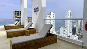 Un immeuble de grande hauteur doté d’un balcon aménagé avec des fauteuils de relaxation en bois et offrant une vue sur la ville.
