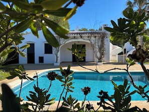 Una piscina rodeada de follaje y un edificio blanco con tejado de baldosas.