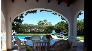Ett takförsett utomhusområde med sittplatser, och en pool syns i bakgrunden.