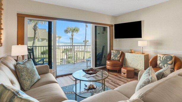 Una sala de estar con un sofá, sillones, una mesa de centro, un televisor y un balcón con vistas a palmeras y la playa.