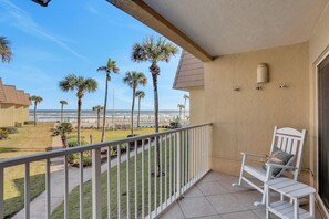 Un balcon avec un fauteuil à bascule blanc, une vue sur les palmiers et la plage, et un bâtiment aux murs de couleur beige et au sol carrelé.