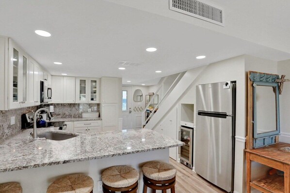 A modern kitchen with white cabinets, a marble countertop, stainless steel appliances, and a wooden dining table with stools.