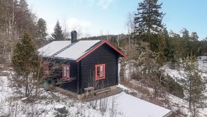 Une petite cabane noire aux bordures rouges et au toit métallique, entourée d’un paysage enneigé et d’arbres.