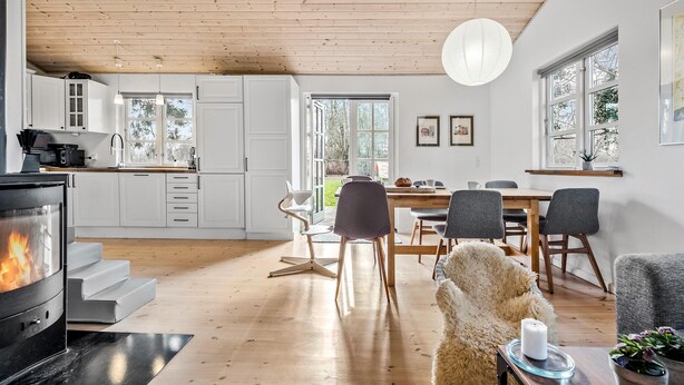 A modern kitchen with white cabinets, a dining area with a wooden table and chairs, and a cozy living space with a fluffy dog and a lit candle. - Stunning home in Holbæk with sauna (Holbæk)