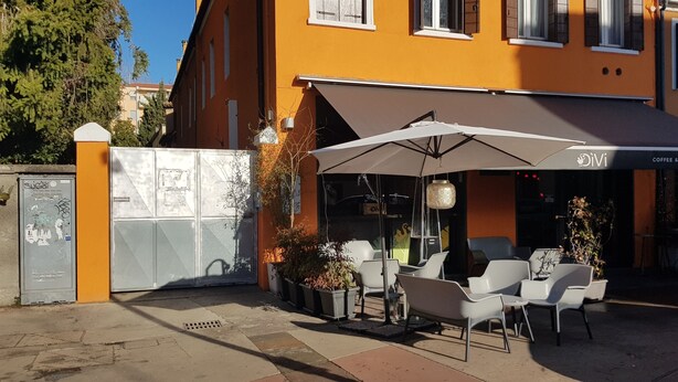 Un espacio de asientos al aire libre con sillas blancas y un paraguas grande, frente a un edificio con un letrero que dice 'Divi Coffee & Co.'