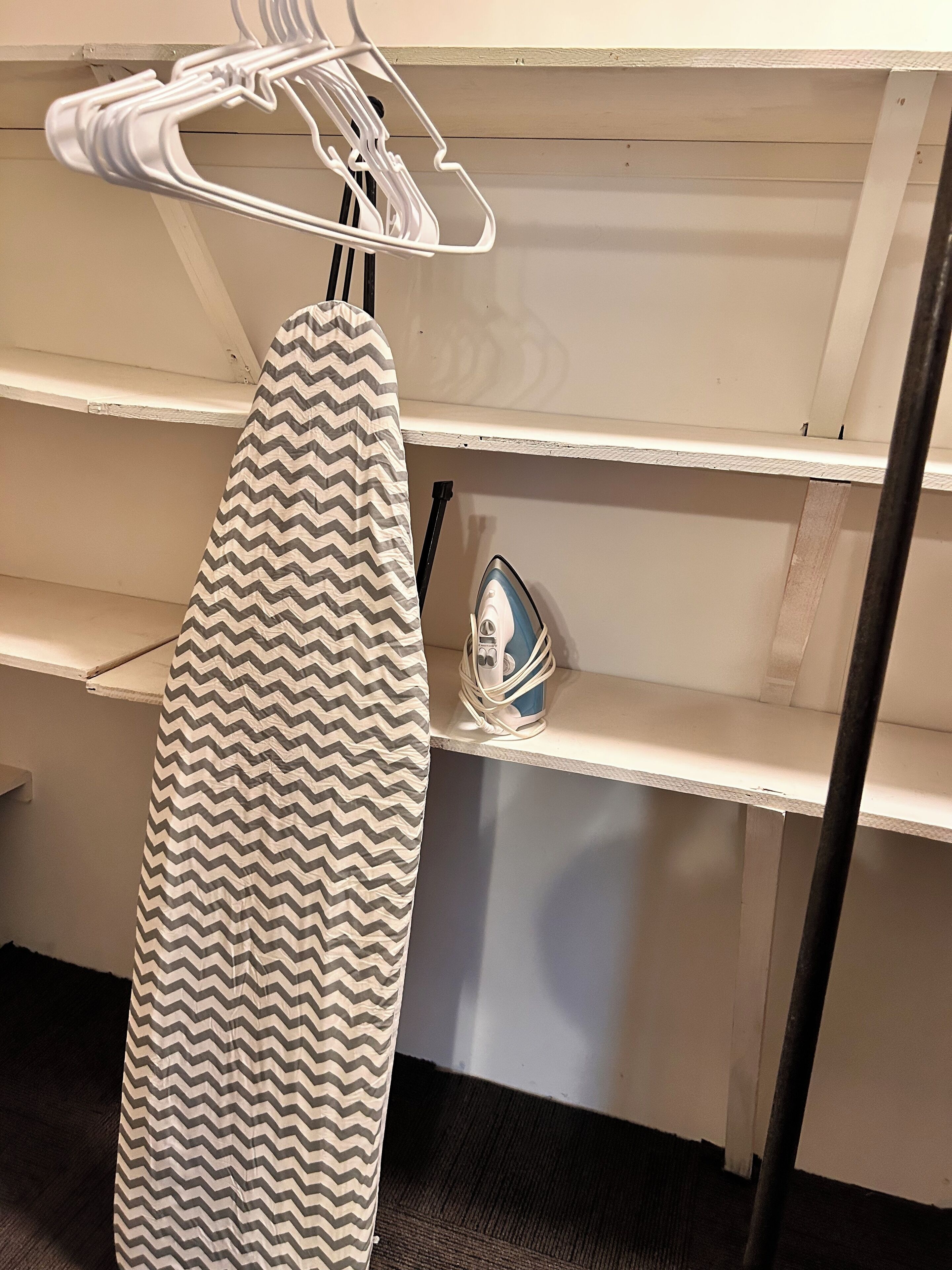 Quarto com um ferro de passar pendurado numa prateleira e um espaço para passar roupa com capa estampada em padrão geométrico.