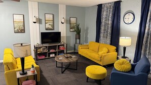 A living room with a yellow sofa, a blue armchair, a coffee table, and a TV on a stand. - Riverside Hideaway, 880sf, 1 bedroom, 2nd story, apartment (Grand Rapids)