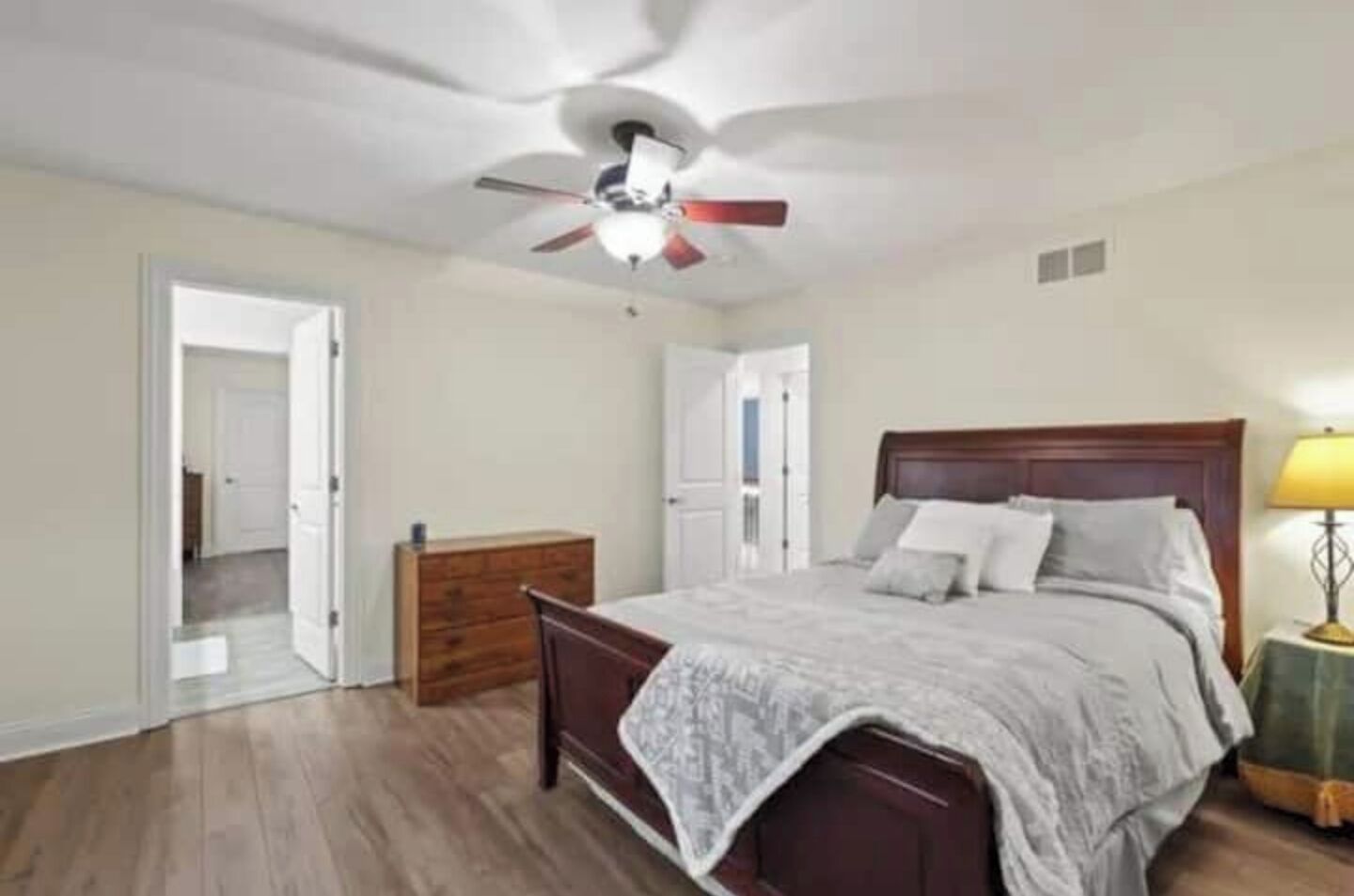 A bedroom with a wooden bed, a nightstand with a lamp, a ceiling fan, and a dresser.