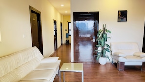 A modern living room with a white sofa, a small glass coffee table, and a white armchair. There is a wooden floor, a door at the end of the room, and a potted plant. - Blue Rocks Suites, Sarjapur Road (Bengaluru)