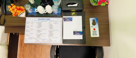 A hotel reception desk with a menu, a phone, a water bottle, a coffee maker, a remote control, a vase of flowers, and a "Please do not disturb" sign.