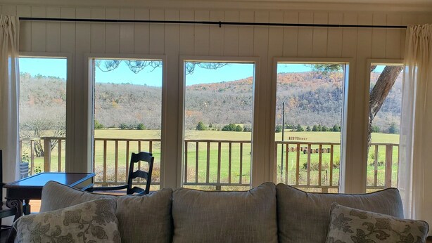 Ein Zimmer mit einer Couch, einem Tisch und einem Stuhl. Große Fenster bieten Blick auf eine Wiese und Hügel.