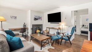 En moderne stue med sofa, lænestol, sofabord, pejs, fladskærms-TV og et skrivebord med lampe.