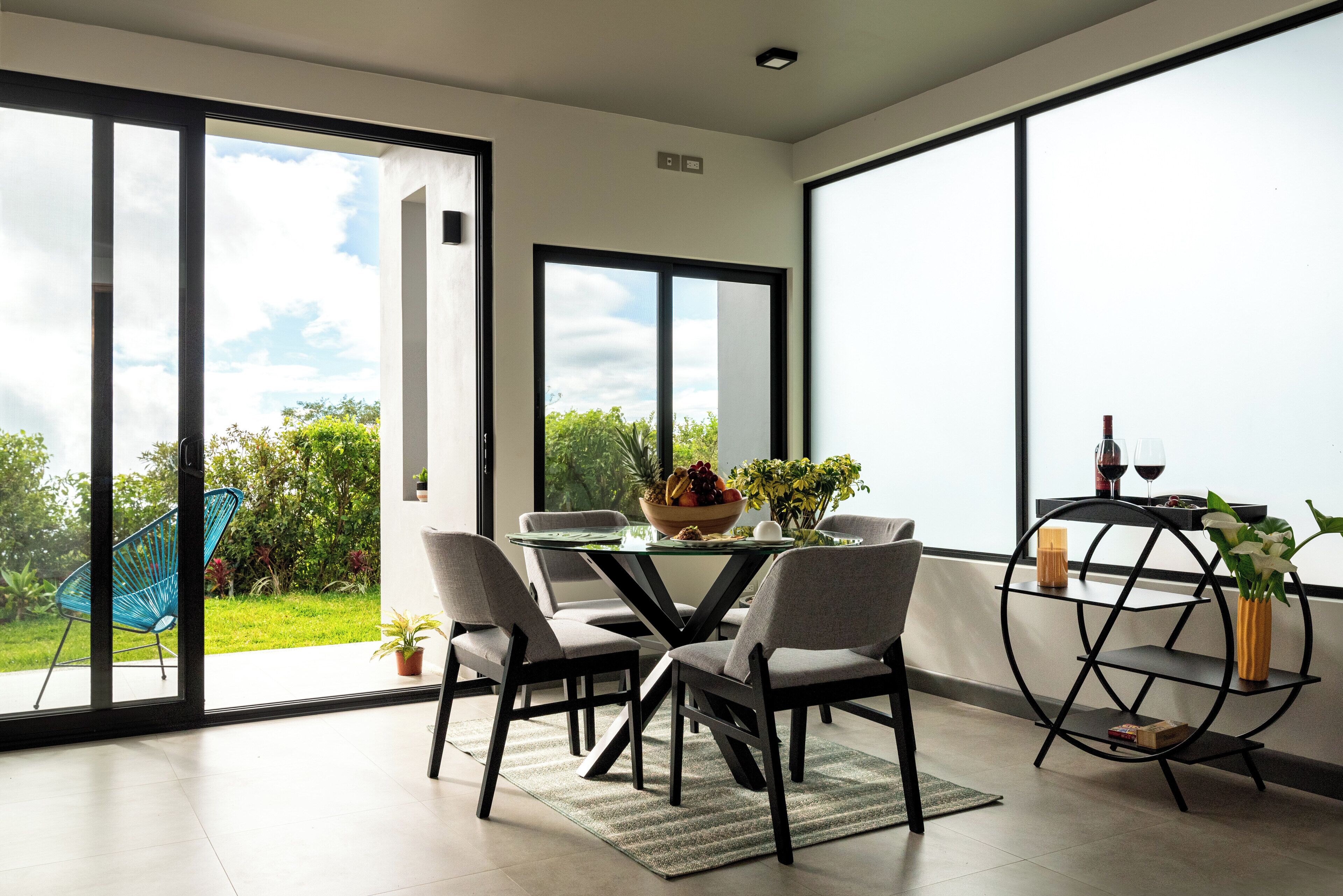 A modern dining area with a glass table, four chairs, and a black metal shelf with a vase.