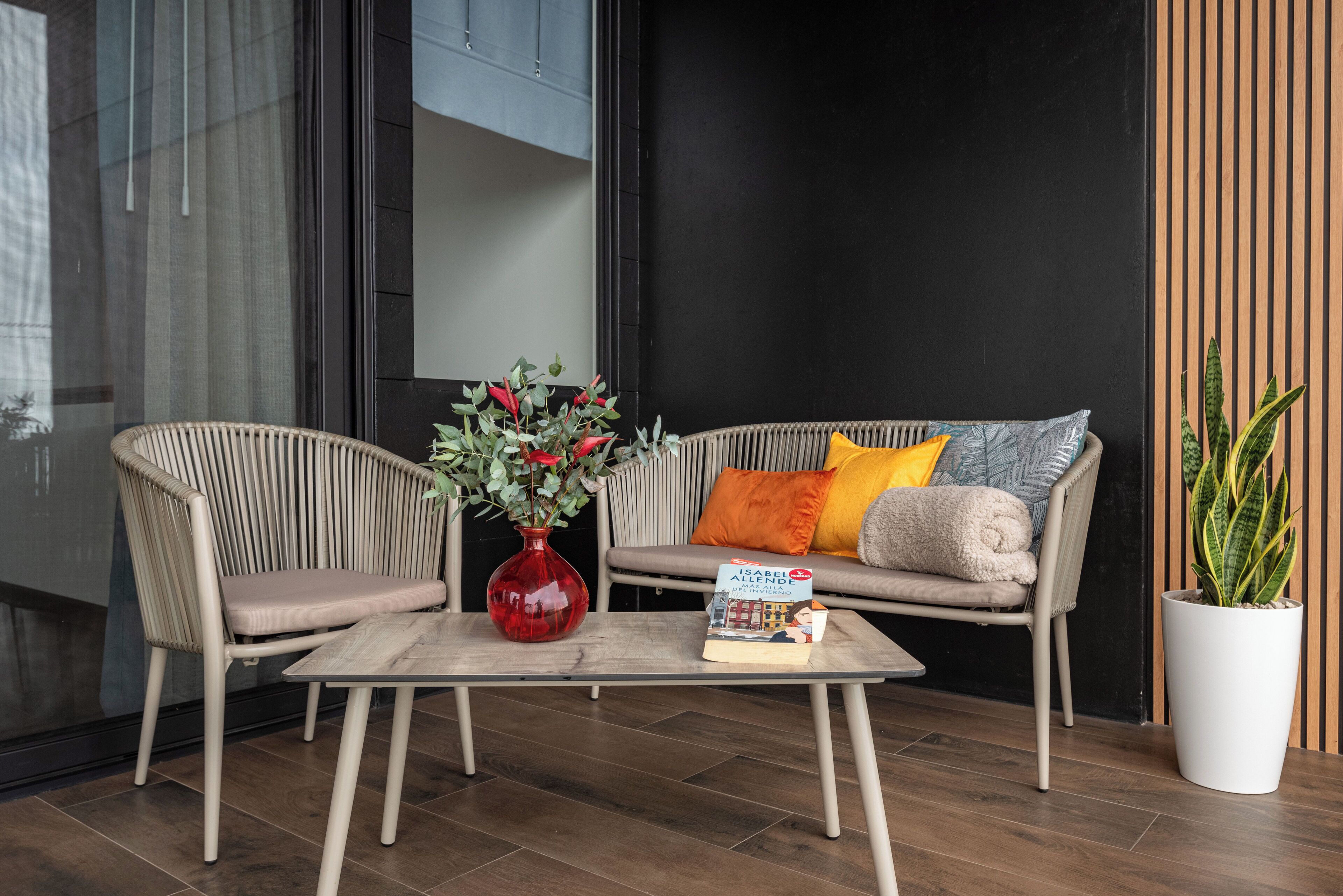 Een moderne zithoek met rieten stoelen, een bank met kussens, een glazen salontafel, een rode vaas met bloemen en een potplant.