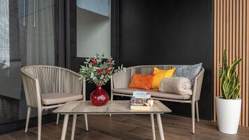 Een moderne zithoek met rieten stoelen, een bank met kussens, een glazen salontafel, een rode vaas met bloemen en een potplant.