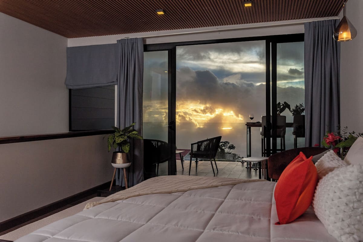 A bedroom with a large bed, a view of the sky through a sliding glass door, and a balcony with chairs and a table.