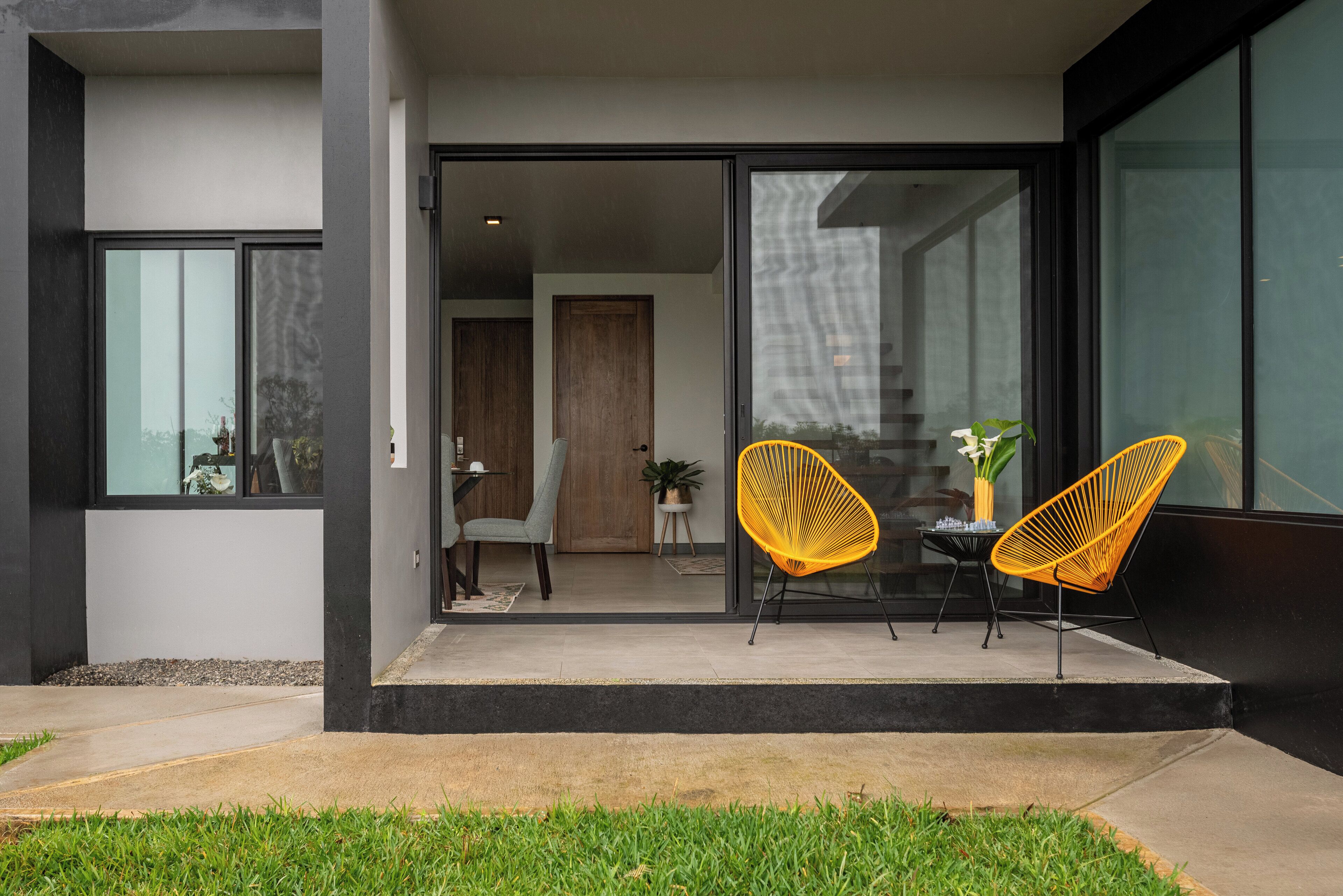 Een modern buitenzitgedeelte met twee gele stoelen en een tafel, een glazen schuifdeur naar een kamer en een kleine tuin.