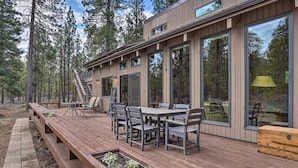 A modern wooden deck with a dining table and chairs, surrounded by large windows and a staircase leading to an upper level. - 5BR. Hot Tub! Full resort access (Sisters)