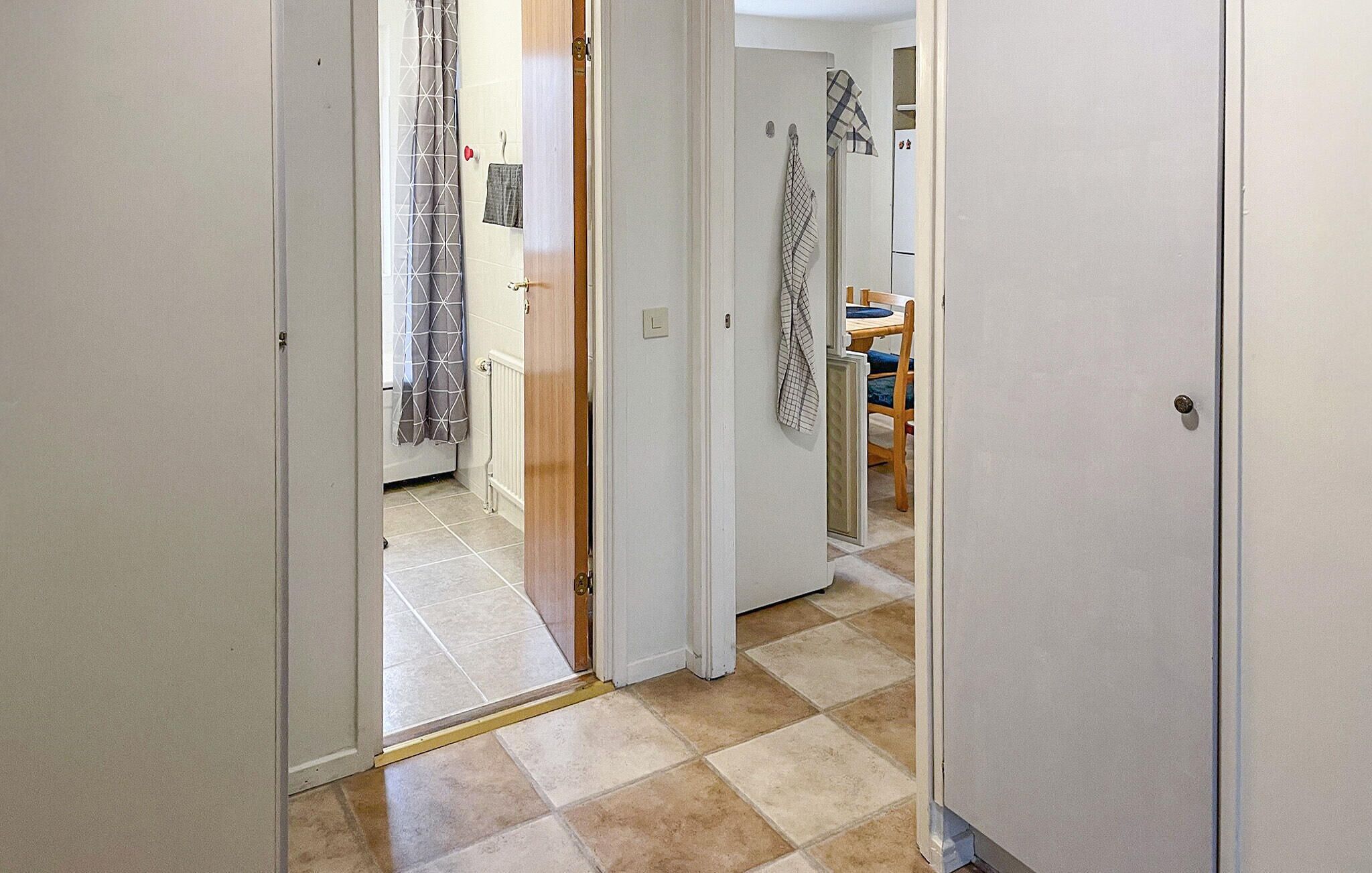 Een hal met betegelde vloer, een deur naar een badkamer met douchegordijn, en een zichtbaar keukenhoekje met een stoel en tafel.