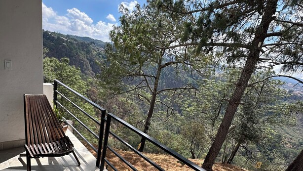 Een balkon met twee houten stoelen en een metalen hekwerk met uitzicht op een bergachtig landschap met bomen.