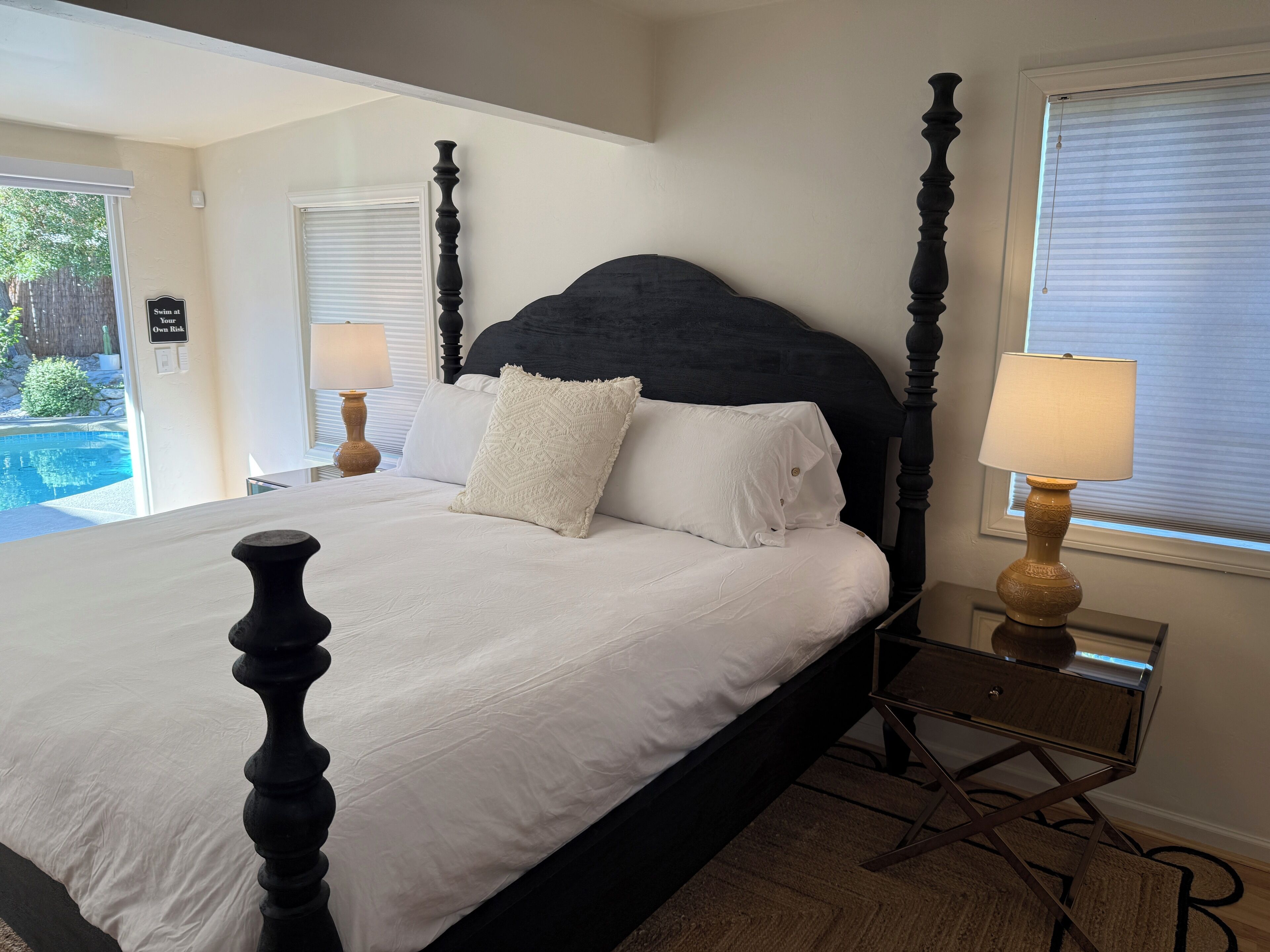 A neatly made bed with a black headboard, white bedding, and a decorative pillow. There is a bedside table with a lamp and a small mirror. The room has a window with blinds, and a sign on the wall that reads "Swim at Your Own Risk."