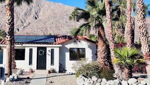 Una casa de un solo piso con techo de tejas, rodeada de palmeras y una ladera rocosa.