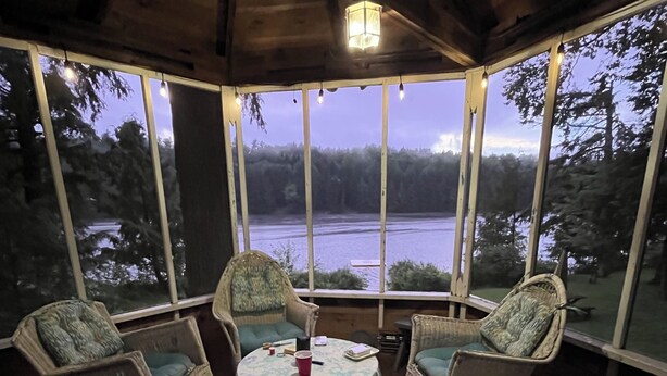 Ein überdachter Außenbereich mit Korbstühlen und einem Tisch, mit Blick auf einen See und bewaldetes Gelände.