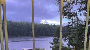 Ein Korbstuhl mit grüner Sitzkissen, ein Tisch mit dekorativem Teller und Blick auf einen See mit Stühlen und einem Picknicktisch.